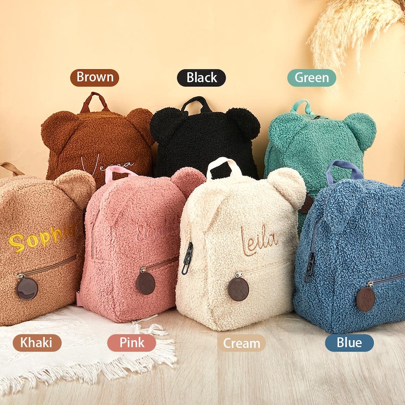K&ouml;nnte beinhalten: Eine Auswahl an Rucks&auml;cken in Teddyb&auml;r-Form in verschiedenen Farben: Braun, Schwarz, Gr&uuml;n, Khaki, Rosa, Creme und Blau. Jeder Rucksack hat einen Rei&szlig;verschluss, einen kleinen runden Anh&auml;nger und gestickte Namen.