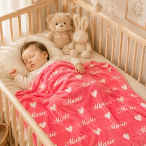 May include: A bright pink plush blanket featuring a repeating pattern of white hearts and the name Marie in a cursive script, covering a sleeping infant in a wooden crib alongside a tan teddy bear and a cream-colored plush rabbit toy.