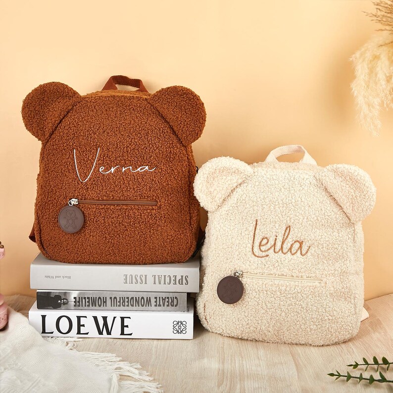 K&ouml;nnte beinhalten: Zwei teddyb&auml;rf&ouml;rmige Rucks&auml;cke. Einer ist braun mit dem gestickten Namen "Verna", der andere ist cremefarben mit dem gestickten Namen "Leila". Beide haben eine Rei&szlig;verschlusstasche und einen runden Anh&auml;nger. Sie stehen auf einem Stapel B&uuml;cher.