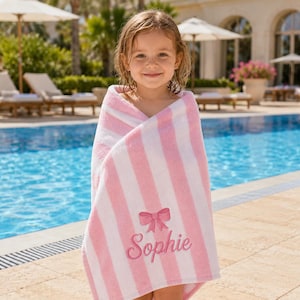 May include: A soft, plush towel featuring vertical light pink and white stripes. The towel is embroidered with a pink bow graphic positioned above the name Sophie, written in a cursive script font. The fabric has a textured, absorbent appearance.