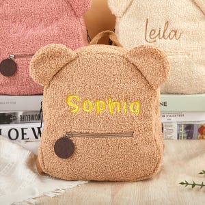 K&ouml;nnte beinhalten: Ein hellbrauner Teddyb&auml;r-Rucksack mit dem Namen "Sophia" in Gelb gestickt. Der Rucksack hat eine Vordertasche mit Rei&szlig;verschluss und ein kleines braunes rundes Etikett. Weitere Rucks&auml;cke in Rosa und Creme sind im Hintergrund zu sehen.