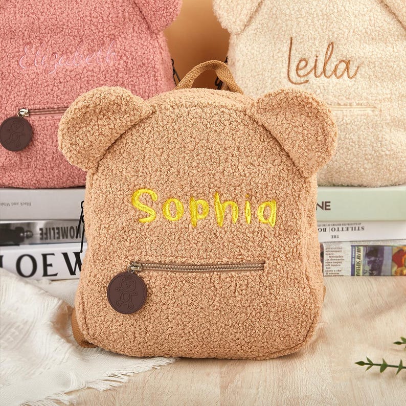 K&ouml;nnte beinhalten: Ein hellbrauner Teddyb&auml;r-Rucksack mit dem Namen "Sophia" in Gelb gestickt. Der Rucksack hat eine Vordertasche mit Rei&szlig;verschluss und ein kleines braunes rundes Etikett. Weitere Rucks&auml;cke in Rosa und Creme sind im Hintergrund zu sehen.