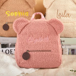 Pu&ograve; includere: Uno zaino rosa a forma di orsetto con il nome "Elizabeth" ricamato sul davanti. Lo zaino ha una tasca con cerniera, una maniglia superiore e orecchie da orso. Sullo sfondo sono visibili altri zaini beige e marroni, ognuno con un nome ricamato.