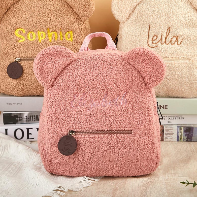 Pu&ograve; includere: Uno zaino rosa a forma di orsetto con il nome "Elizabeth" ricamato sul davanti. Lo zaino ha una tasca con cerniera, una maniglia superiore e orecchie da orso. Sullo sfondo sono visibili altri zaini beige e marroni, ognuno con un nome ricamato.