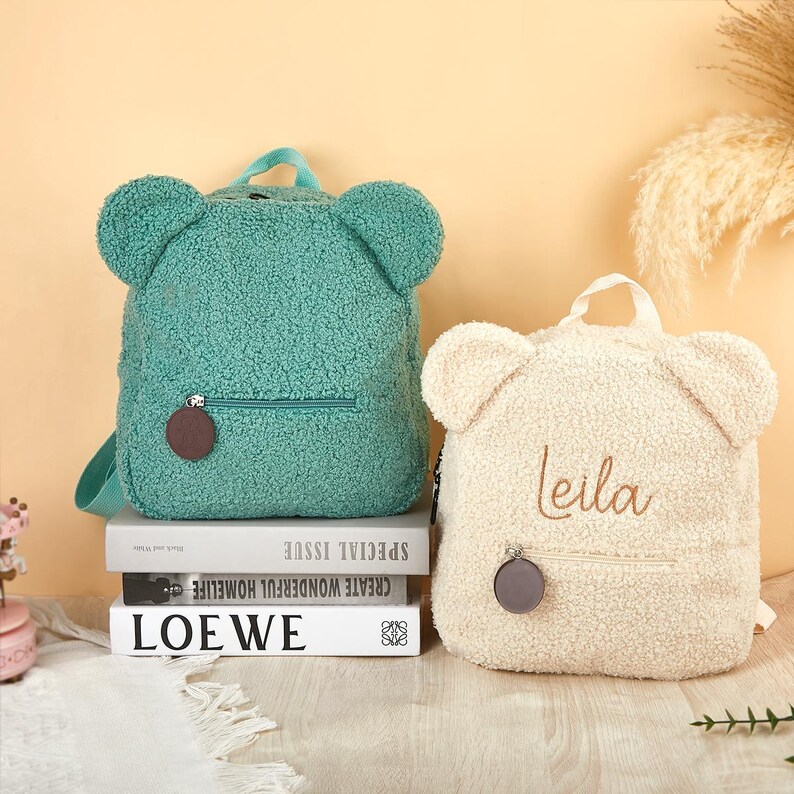 K&ouml;nnte beinhalten: Zwei teddyb&auml;rf&ouml;rmige Rucks&auml;cke. Einer ist t&uuml;rkis, der andere cremefarben mit dem Namen "leila" darauf gestickt. Beide haben eine Rei&szlig;verschlusstasche und einen runden, braunen Rei&szlig;verschlussanh&auml;nger. Sie liegen auf B&uuml;chern.
