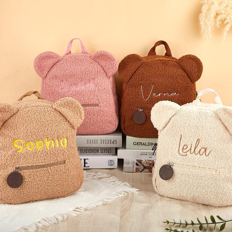 Pu&ograve; includere: Quattro zaini a forma di orsetto nei colori rosa, marrone, beige e crema. Ogni zaino ha una tasca con cerniera, un manico e nomi ricamati: Sophia, Verna e Leila. Gli zaini sono appoggiati sopra dei libri.
