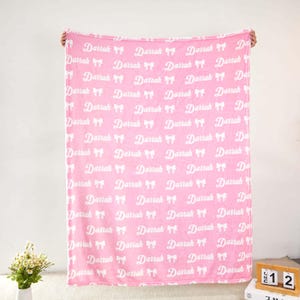 May include: A soft pink fleece blanket featuring a repeating white pattern of the name Darrah in a cursive script, interspersed with small white bow icons. The blanket is held up vertically, displaying the full print across its entire surface.