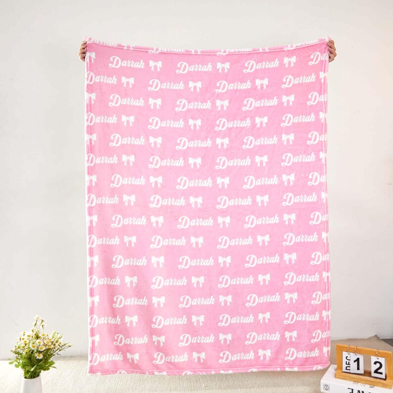 May include: A soft pink fleece blanket featuring a repeating white pattern of the name Darrah in a cursive script, interspersed with small white bow icons. The blanket is held up vertically, displaying the full print across its entire surface.