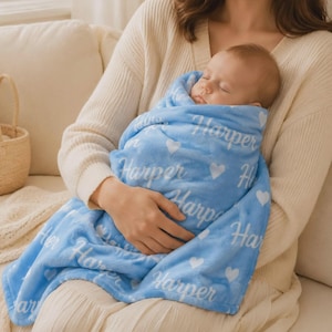 May include: A soft light blue baby blanket featuring a repeating pattern of white hearts and the name Harper printed in a white cursive script. The blanket is wrapped around a sleeping infant, showcasing the plush texture and the personalized design.