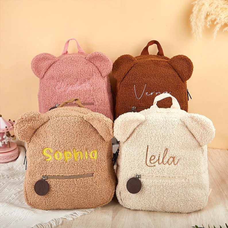 K&ouml;nnte beinhalten: Vier teddyb&auml;rf&ouml;rmige Rucks&auml;cke in Rosa, Braun, Beige und Creme. Jeder Rucksack hat eine Rei&szlig;verschlusstasche, einen oberen Griff und gestickte Namen: "Sophia", "Verna" und "Leila". Die Rucks&auml;cke haben B&auml;renohren.