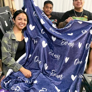 May include: A navy blue blanket with white hearts and the name "Evan" in cursive script. The blanket is held by three people, showcasing its size and design. The blanket appears soft and cozy.