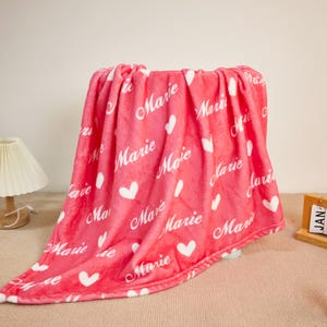Op de afbeelding: Een roze fleece deken met de naam "Marie" in witte letters en witte hartvormen. De deken is over een stoel gedrapeerd, wat de zachte textuur en het ontwerp benadrukt. Een gezellig woondecoratie-item.