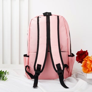 May include: A light pink backpack featuring black trim and black adjustable shoulder straps. The bag includes two side pockets and a top handle, showcasing a simple design with a main compartment and a front zippered pocket for storage.