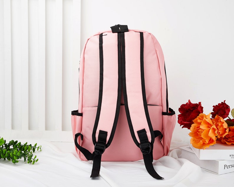 May include: A light pink backpack featuring black trim and black adjustable shoulder straps. The bag includes two side pockets and a top handle, showcasing a simple design with a main compartment and a front zippered pocket for storage.