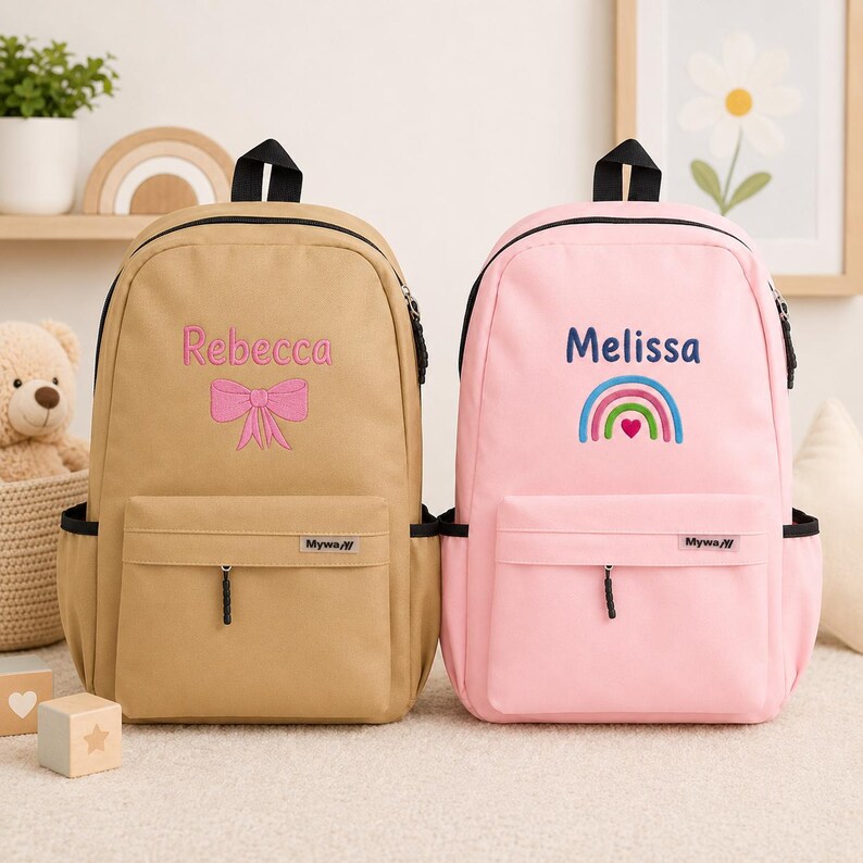 May include: Two backpacks with front zippered pockets and side pouches. The tan backpack features the embroidered name Rebecca and a pink bow. The pink backpack features the embroidered name Melissa and a rainbow with a heart. Both have Mywa labels.