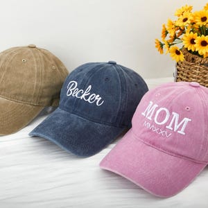 Może przedstawiać: Trzy czapki bejsbolowe w różnych kolorach z haftowanym tekstem. Pierwsza czapka jest brązowa bez tekstu. Druga czapka jest niebieska z haftowanym tekstem "Becker" w kolorze białym. Trzecia czapka jest różowa z haftowanym tekstem "MOM MMXXV" w kolorze białym.