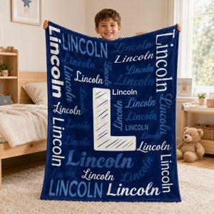 May include: A dark blue plush blanket featuring the name Lincoln repeated in various white and light blue fonts, with a large white letter L centered on the fabric. The text is oriented in multiple directions across the soft material.