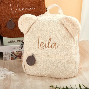 Pu&ograve; includere: Uno zaino a forma di orsetto color crema con il nome "Leila" ricamato con filo marrone chiaro. Lo zaino ha le orecchie da orso e una piccola tasca frontale con cerniera. Un ciondolo circolare marrone pende dalla cerniera.