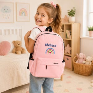 May include: A light pink backpack featuring the name Melissa embroidered in blue text above a colorful rainbow graphic with a small red heart at the center. The bag includes a front zippered pocket, black straps, and side mesh pockets for storage.