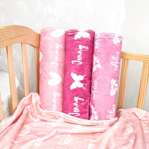May include: Three rolled plush blankets in shades of pink, featuring white printed designs. The left blanket shows hearts and the name Jenny, the center shows butterflies and the name Jenny, and the right shows bows and the name Alice.