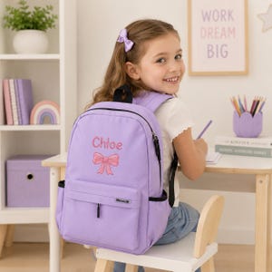 May include: A light purple backpack featuring the embroidered name Chloe in pink script above a pink bow graphic. The bag includes a front zippered pocket with a small rectangular label, black trim, side pockets, and adjustable shoulder straps.