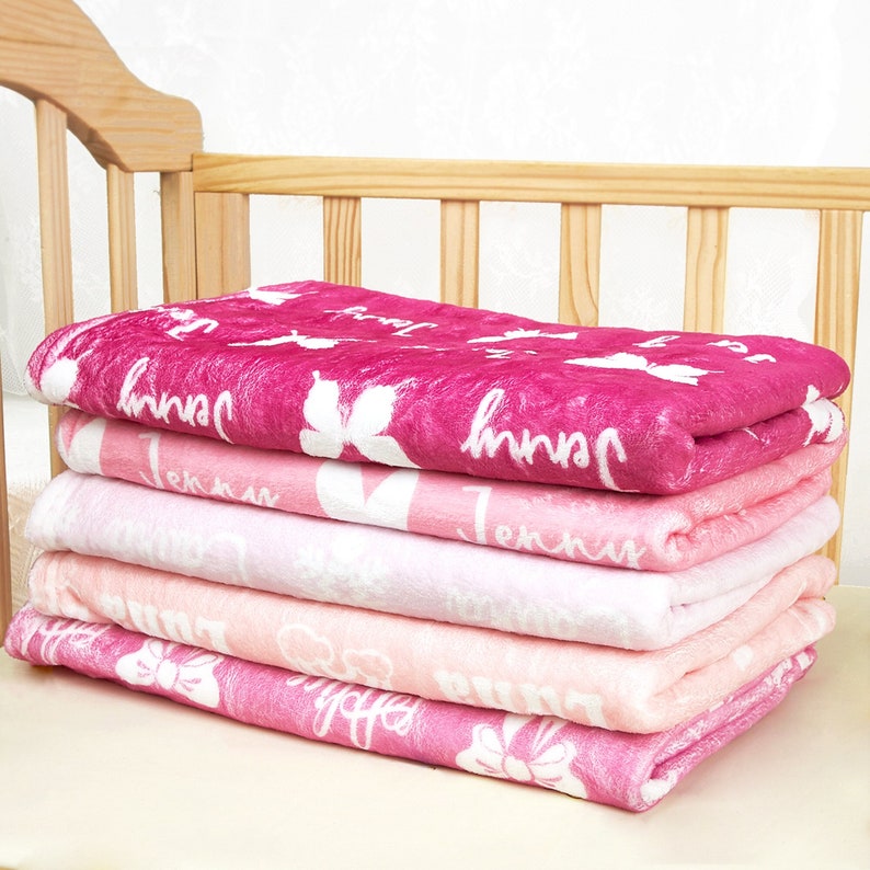 May include: A stack of five folded plush blankets in varying shades of pink, ranging from deep magenta to light pastel pink. Each blanket features a repeating white pattern of butterflies, bows, and the name Jenny printed in a cursive script.