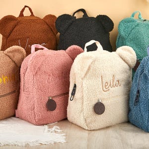 Pu&ograve; includere: Una collezione di zaini a forma di orsetto in vari colori, tra cui marrone, nero, rosa e crema. Ogni zaino presenta una tasca frontale con cerniera, una maniglia superiore e spallacci regolabili. Alcuni zaini hanno nomi ricamati.