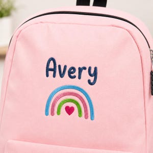 May include: A light pink backpack featuring the embroidered name Avery in dark blue text. Below the name is a colorful embroidered rainbow design with blue, pink, and green arches, centered with a small, solid pink heart at the base.