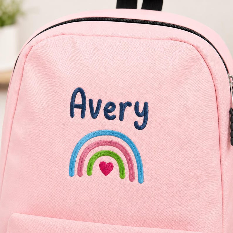 May include: A light pink backpack featuring the embroidered name Avery in dark blue text. Below the name is a colorful embroidered rainbow design with blue, pink, and green arches, centered with a small, solid pink heart at the base.