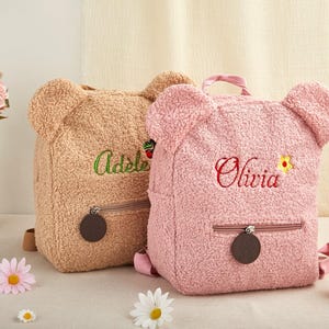 May include: Two teddy bear-shaped backpacks. One is tan with the name "Adele" embroidered in green. The other is pink with "Olivia" embroidered in red. Both have zippered pockets and bear ears.