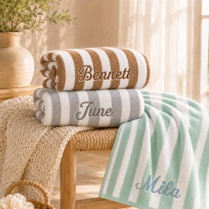 May include: Three folded towels with vertical stripes in brown, gray, and light green. Each towel features embroidered names in cursive script: Bennett on the brown towel, June on the gray towel, and Mila on the light green towel.