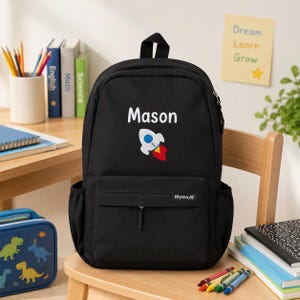 May include: A black backpack featuring the name Mason in white text above a graphic of a white, blue, and red rocket ship. The bag has a front zippered pocket with a small Myway brand tag and side pockets, constructed from a solid black fabric.