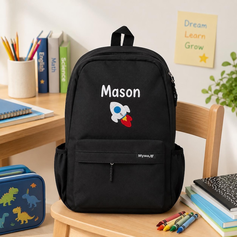 May include: A black backpack featuring the name Mason in white text above a graphic of a white, blue, and red rocket ship. The bag has a front zippered pocket with a small Myway brand tag and side pockets, constructed from a solid black fabric.