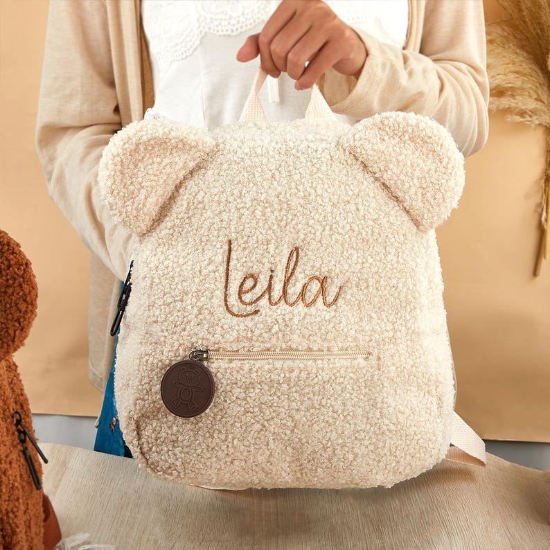 K&ouml;nnte beinhalten: Ein cremefarbener Rucksack mit Teddyb&auml;r-Design, mit zwei gro&szlig;en Ohren oben. Der Name "Leila" ist in einer Schreibschrift gestickt. Der Rucksack hat eine Rei&szlig;verschluss-Vordertasche und einen kleinen, runden Anh&auml;nger.
