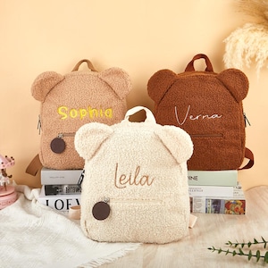 K&ouml;nnte beinhalten: Drei teddyb&auml;rf&ouml;rmige Rucks&auml;cke in den Farben Beige, Braun und Creme. Jeder Rucksack hat einen Rei&szlig;verschluss, verstellbare Tr&auml;ger und einen kleinen runden Anh&auml;nger. Die Namen "Sophia", "Verna" und "Leila" sind aufgestickt.