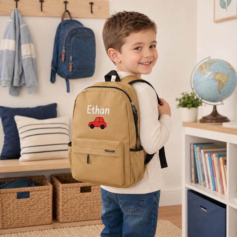 May include: A tan backpack featuring a small red car graphic and the name Ethan printed in white text. The backpack has a main compartment, a front zippered pocket, black straps, and side mesh pockets, designed for carrying school supplies.