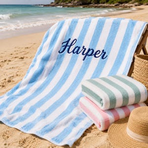 May include: A large beach towel with light blue and white vertical stripes featuring the name Harper embroidered in dark blue script. Two folded beach towels with green and white or pink and white stripes are stacked next to a straw hat.