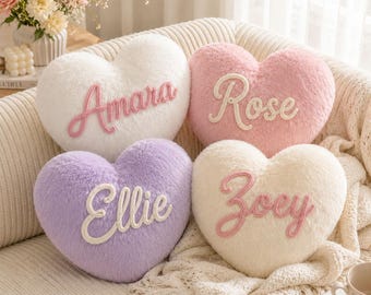 May include: Four heart-shaped plush cushions with soft, fuzzy textures. The white cushion features the name Amara in pink, the pink cushion has Rose in white, the lavender cushion displays Ellie in white, and the cream cushion shows Zoey in pink.