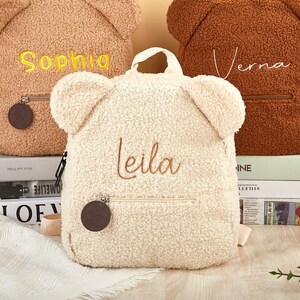 K&ouml;nnte beinhalten: Cremefarbener Teddyb&auml;r-Rucksack mit dem Namen "Leila" in braunem Faden gestickt. Der Rucksack hat eine Rei&szlig;verschlusstasche und einen kleinen Griff. Zwei weitere Rucks&auml;cke sind sichtbar, einer in Beige mit "Sophia" und einer in Braun mit "Verna".