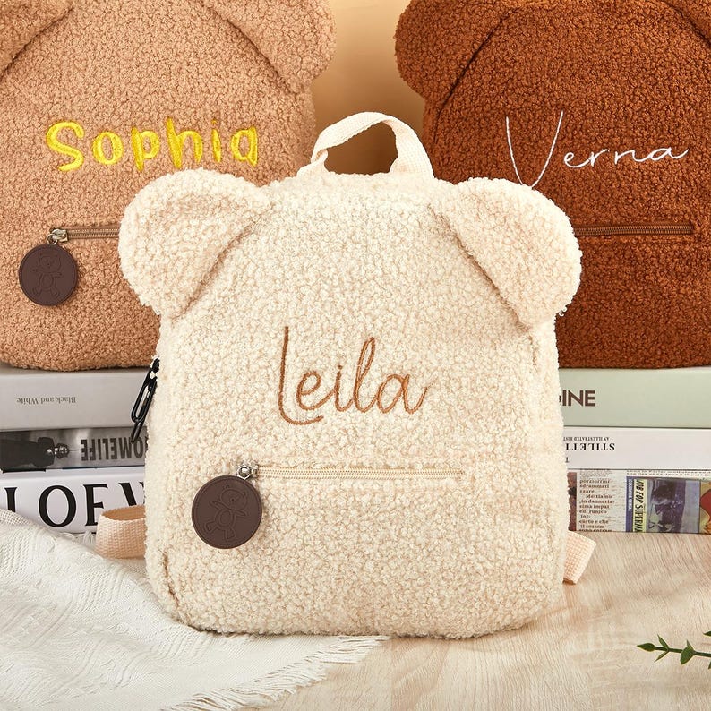 K&ouml;nnte beinhalten: Cremefarbener Teddyb&auml;r-Rucksack mit dem Namen "Leila" in braunem Faden gestickt. Der Rucksack hat eine Rei&szlig;verschlusstasche und einen kleinen Griff. Zwei weitere Rucks&auml;cke sind sichtbar, einer in Beige mit "Sophia" und einer in Braun mit "Verna".