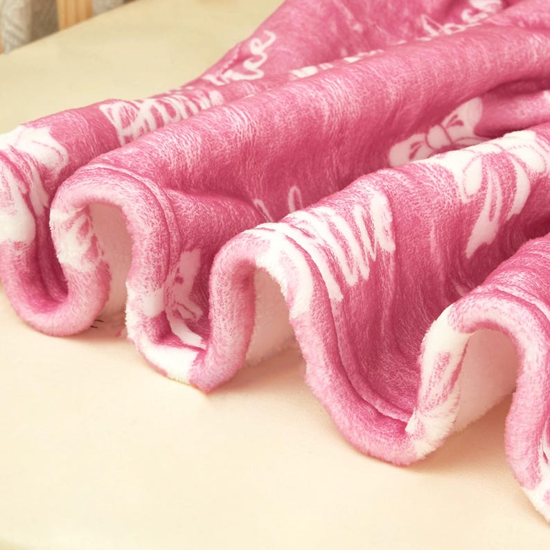 May include: A soft, plush blanket in a vibrant shade of pink featuring a repeating white pattern of bows and cursive script text. The fabric has a fuzzy, textured surface with deep, flowing folds that highlight the material's thickness and softness.