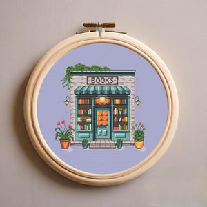 Op de afbeelding: Een kruissteekborduurwerk in een ronde houten ring met een boekenwinkel op een lichtpaarse achtergrond. De winkel heeft een groenblauwe deur, planken vol kleurrijke boeken, een gestreepte luifel en potplanten aan weerszijden.