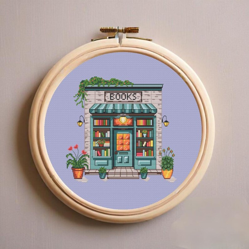 Op de afbeelding: Een kruissteekborduurwerk in een ronde houten ring met een boekenwinkel op een lichtpaarse achtergrond. De winkel heeft een groenblauwe deur, planken vol kleurrijke boeken, een gestreepte luifel en potplanten aan weerszijden.