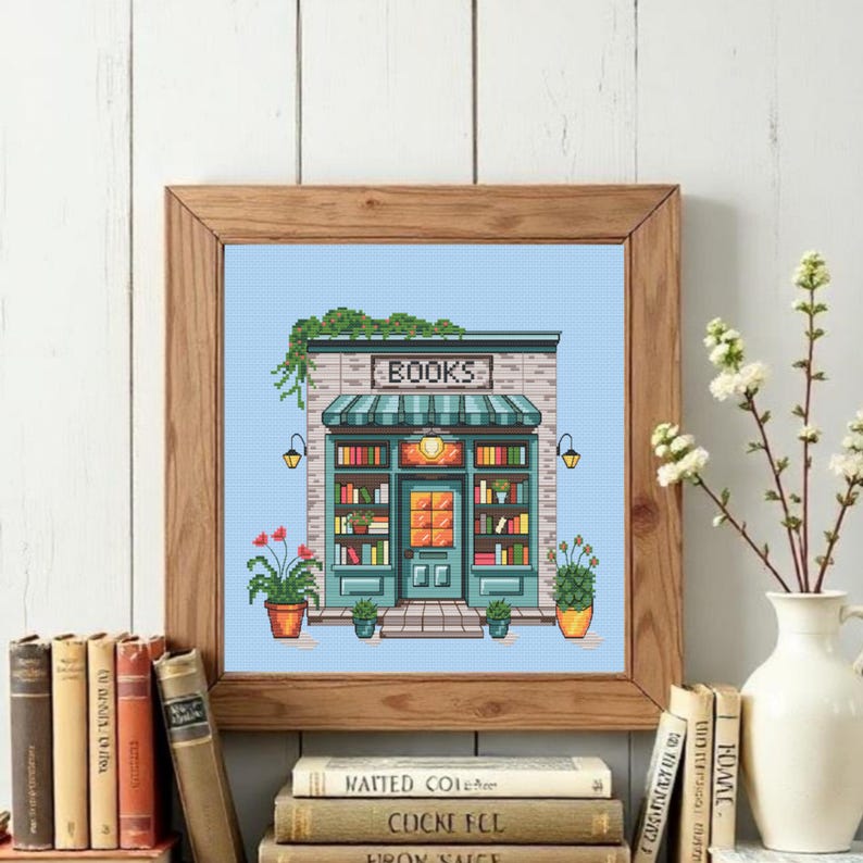 Op de afbeelding: Een ingelijst kruissteekpatroon met een boekenwinkelgevel en het woord BOOKS boven een blauwgroene luifel. Het ontwerp toont planken vol kleurrijke boeken, een centrale deur, twee wandlampen en potplanten op een lichtblauwe achtergrond.