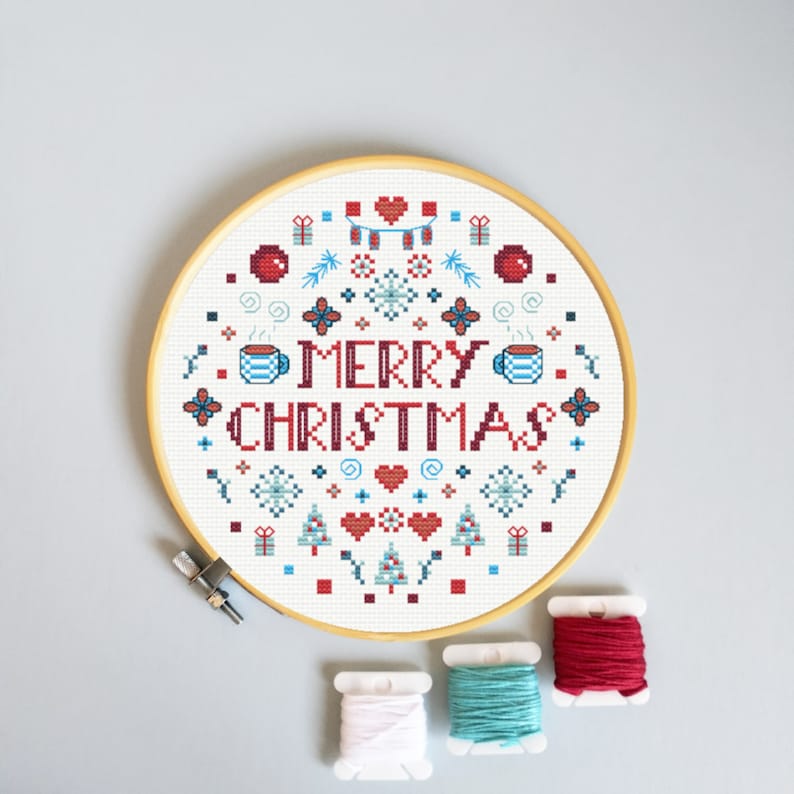 May include: A white cross-stitch embroidery hoop with a red and blue design that says "Merry Christmas". The design includes snowflakes, hearts, Christmas trees, and presents.