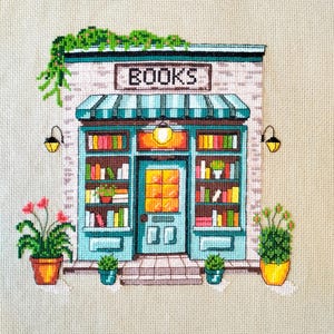 Op de afbeelding: Een kruissteekborduurwerk op beige stof met de voorgevel van een boekwinkel met een blauwgroen frame, een gestreepte luifel en het woord BOOKS boven de deur. De winkel heeft planken vol kleurrijke boeken, twee wandlampen en planten.
