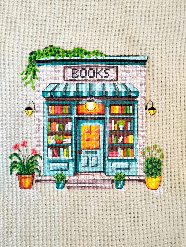 Op de afbeelding: Een kruissteekborduurwerk op beige stof met de voorgevel van een boekwinkel met een blauwgroen frame, een gestreepte luifel en het woord BOOKS boven de deur. De winkel heeft planken vol kleurrijke boeken, twee wandlampen en planten.