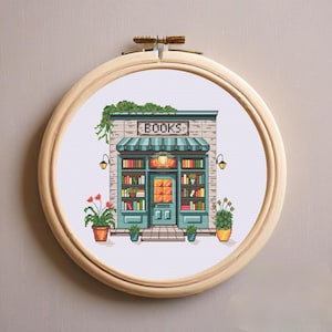 Op de afbeelding: Een kruissteekborduurwerk in een ronde houten ring met een boekenwinkelgevel, een blauwgroene luifel, een bord met de tekst BOOKS, planken vol kleurrijke boeken, een centrale deur en potplanten aan weerszijden van de ingang.