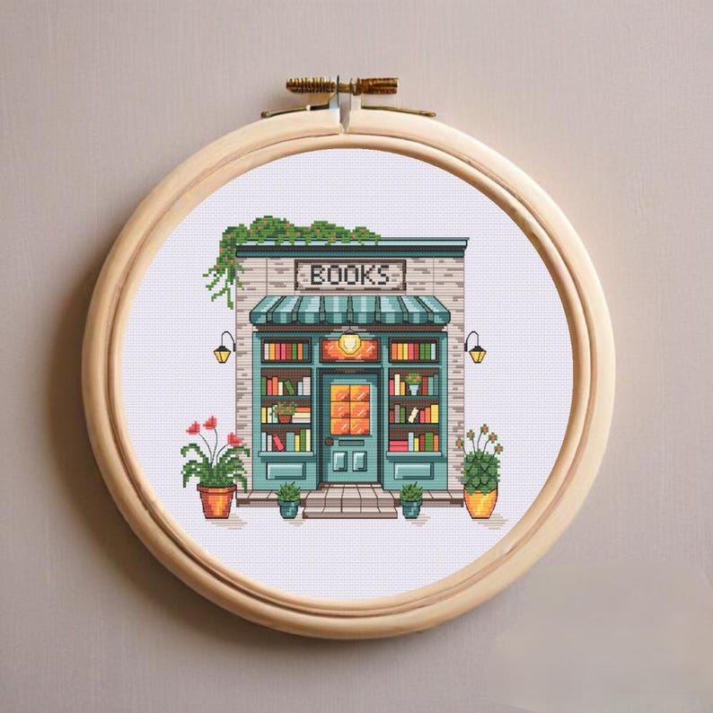 Op de afbeelding: Een kruissteekborduurwerk in een ronde houten ring met een boekenwinkelgevel, een blauwgroene luifel, een bord met de tekst BOOKS, planken vol kleurrijke boeken, een centrale deur en potplanten aan weerszijden van de ingang.