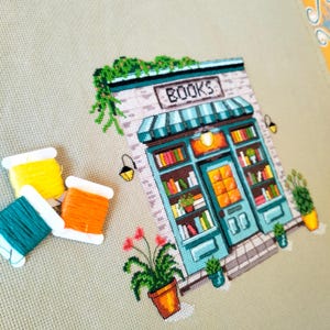 Op de afbeelding: Een kruissteekborduurwerk op lichtbeige stof met een boekwinkel met een blauwgroene pui, een gestreepte luifel en het woord BOOKS boven de deur. Drie klossen borduurgaren in geel, blauwgroen en oranje liggen aan de linkerkant.